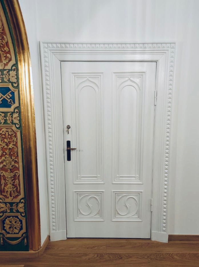 Białe, zdobione drzwi wewnętrzne z czarną klamką i kluczem, z dekoracyjną listwą przypodłogową i fragmentem zdobionej ściany w tle. Parkiet na podłodze.