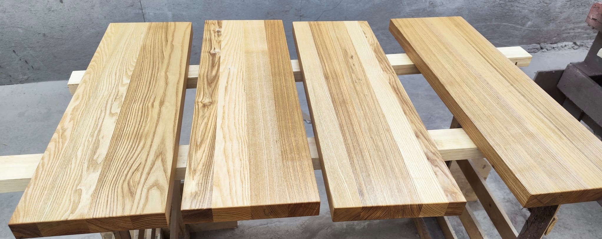 Cztery drewniane blaty z wyraźnym usłojeniem, leżące na drewnianej konstrukcji. Blaty są jasne, w odcieniach beżu i brązu, prawdopodobnie po lakierowaniu lub olejowaniu.