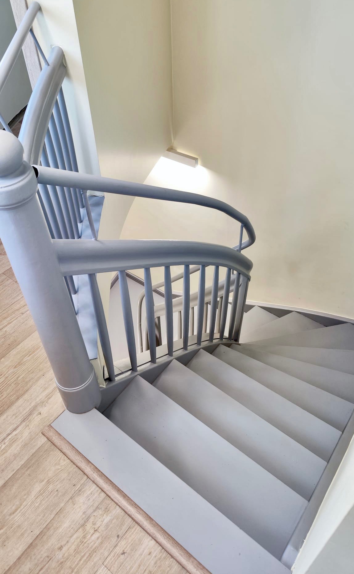 Spiralne schody w odcieniach szarości, widok z góry, z drewnianą podłogą na dole i kremową ścianą w tle, oświetlone delikatnym światłem.