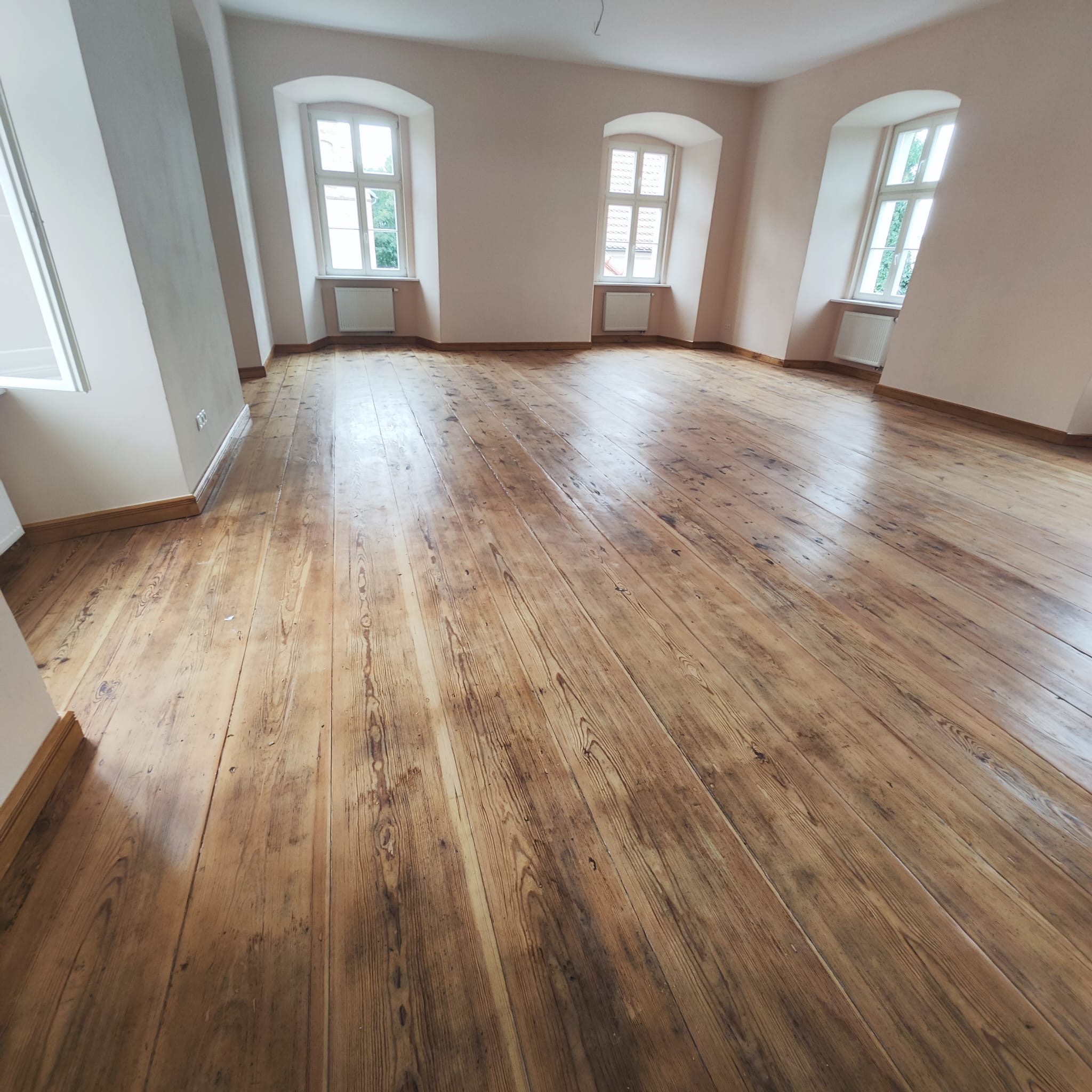 Jasne, przestronne wnętrze z odnowioną, drewnianą podłogą z widocznymi słojami. Trzy okna z białymi ramami wpuszczają naturalne światło. Listwy przypodłogowe w kolorze drewna.