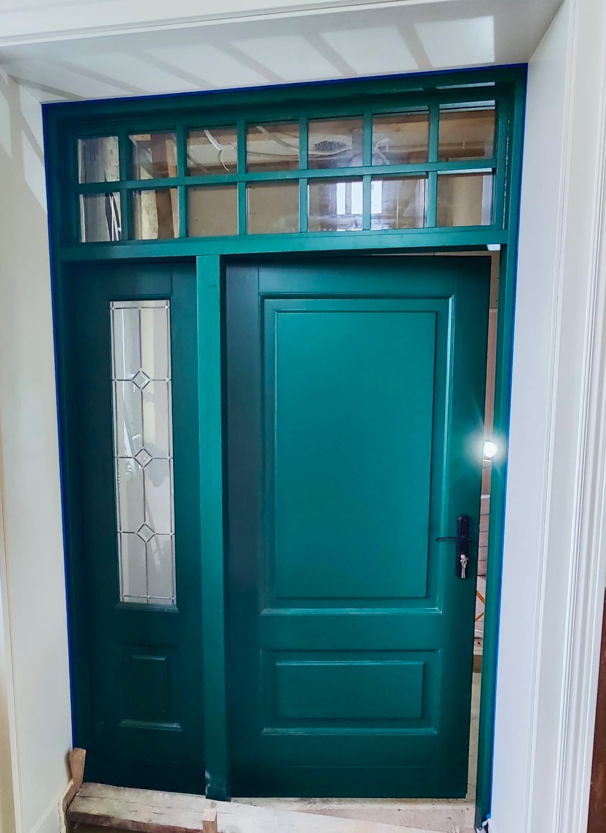 Zielone, odnowione drzwi wejściowe z szybą i nadświetlem, zabezpieczone taśmą malarską. Widoczne ślady prac wykończeniowych.
