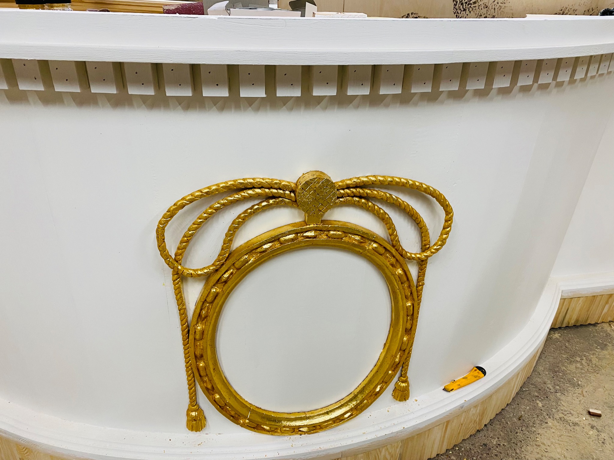 Biała, zaokrąglona konstrukcja z dekoracyjnym, złotym ornamentem w kształcie ramy i sznura. Nóż introligatorski leży obok. Detal wykończenia wnętrza.