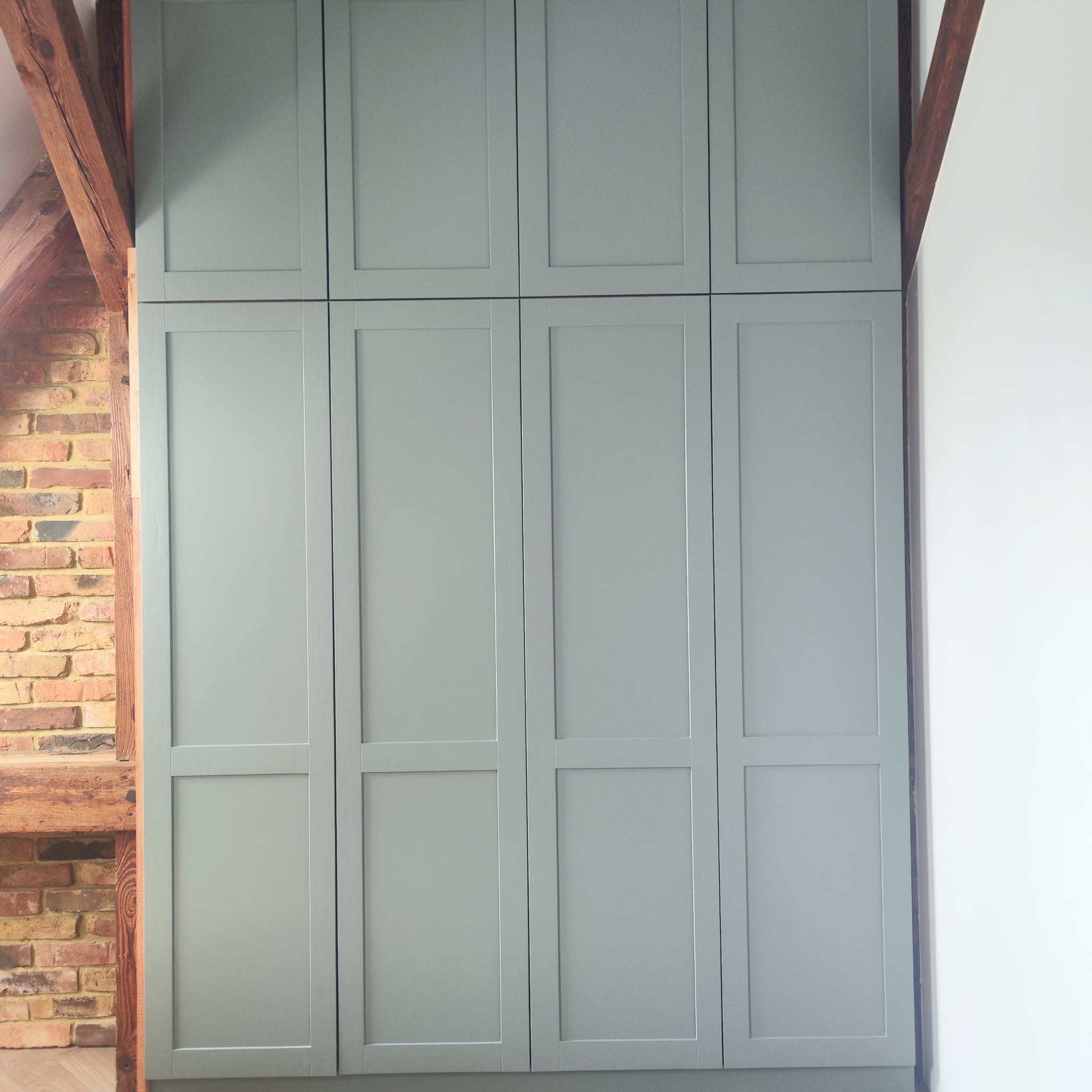 Szara zabudowa szafy wnękowej z drewnianą konstrukcją stropu i ceglaną ścianą w tle. Widok na fronty szafy w szaro-zielonym odcieniu.