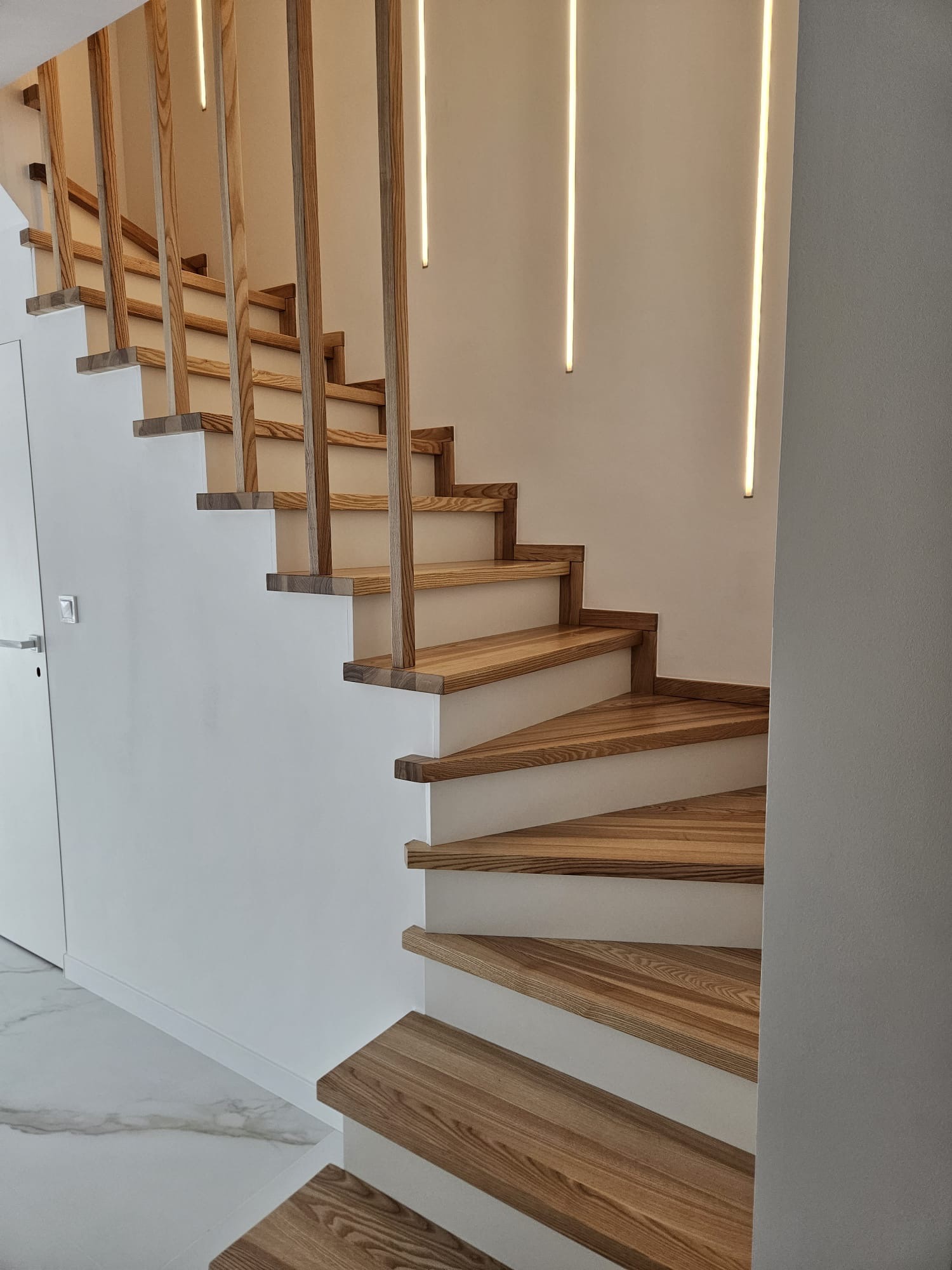 Drewniane schody z jasnego drewna z minimalistyczną balustradą i oświetleniem LED na ścianie. Nowoczesny design, białe ściany i marmurowa podłoga.