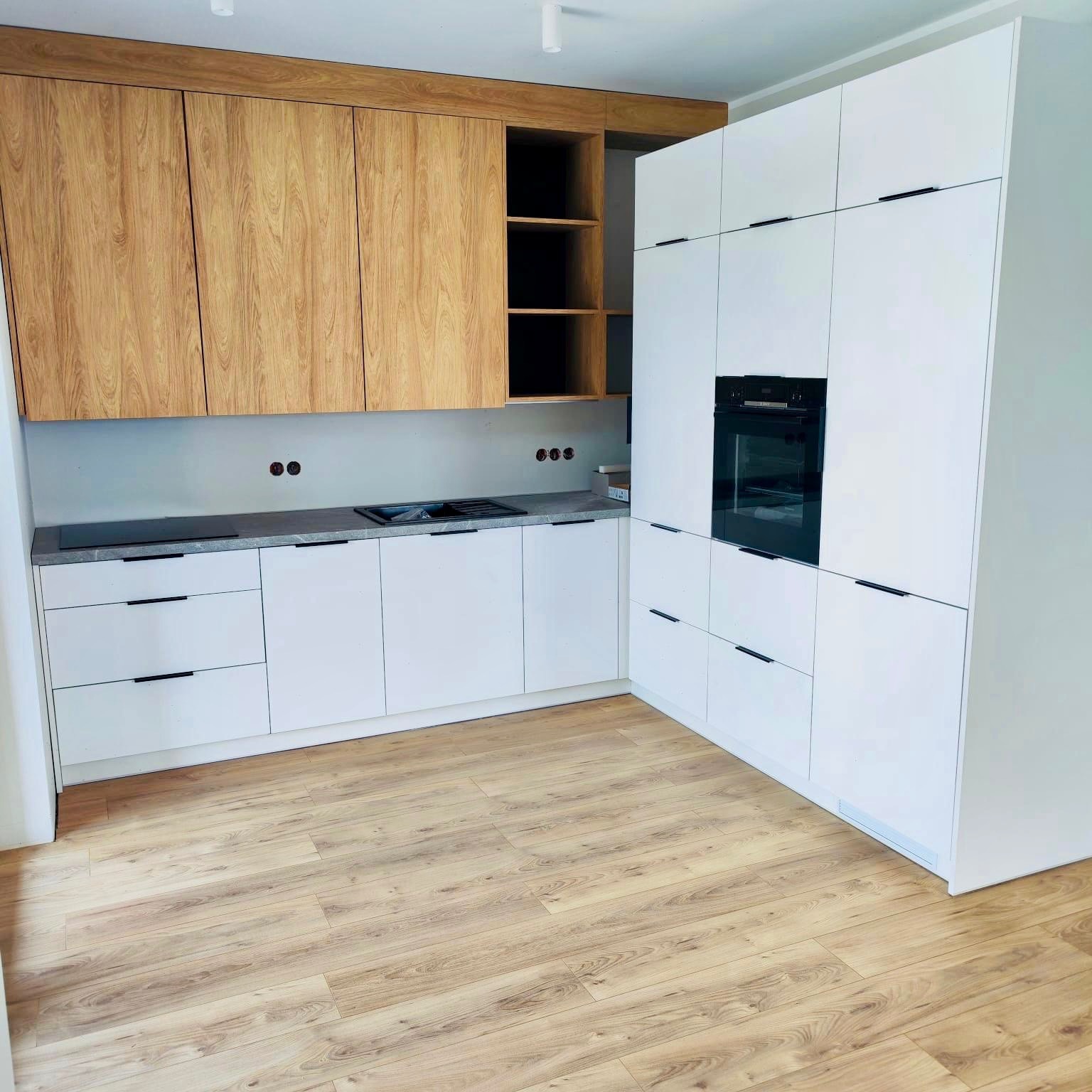 Nowoczesna kuchnia z białymi szafkami i drewnianymi akcentami. Widoczny piekarnik w zabudowie i drewniana podłoga. Minimalistyczny design, jasne wnętrze.