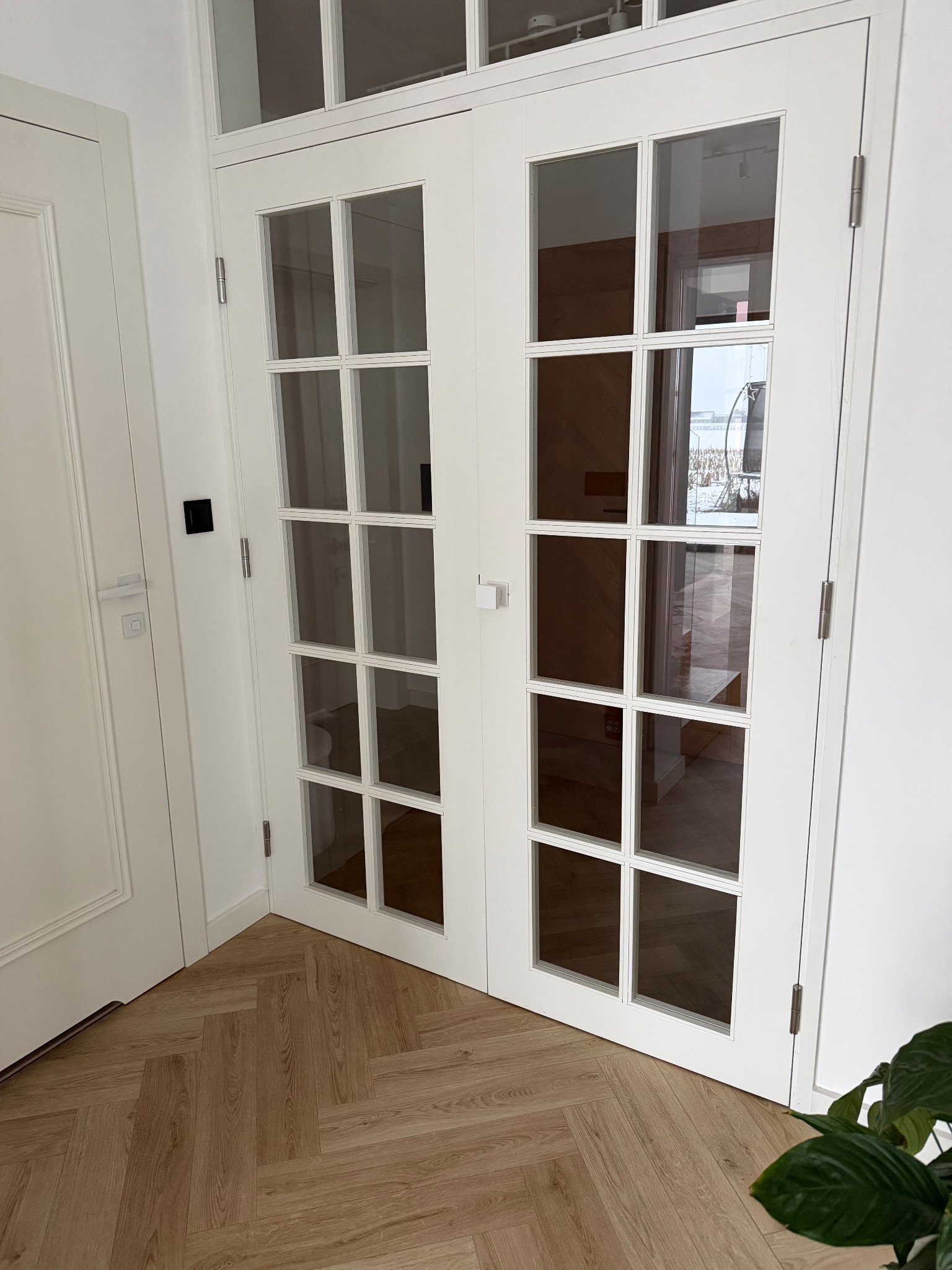 Białe, przeszklone drzwi dwuskrzydłowe z widoczną podłogą w jodełkę. Widok na wnętrze przez szyby. Minimalistyczny design.