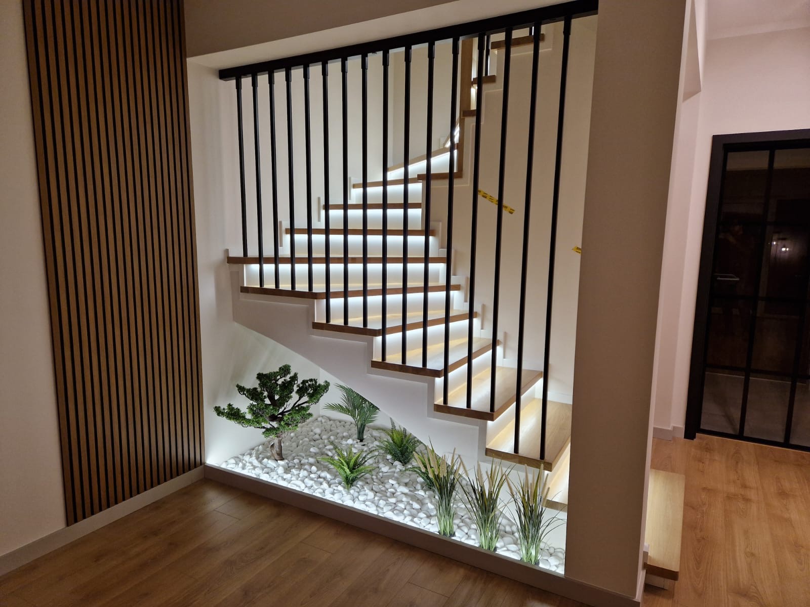 Nowoczesne schody drewniane z podświetleniem LED i dekoracyjnym ogrodem z białych kamieni. Czarna balustrada i drewniana ściana lamelowa.