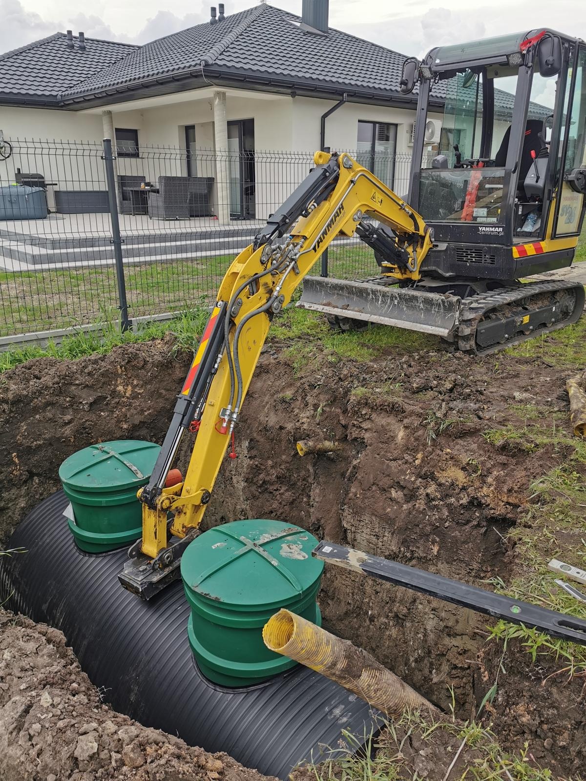 Żółta koparka umieszcza elementy czarnej instalacji z zielonymi pokrywami w wykopie ziemnym na tle domu z szarą dachówką i ogrodzenia.