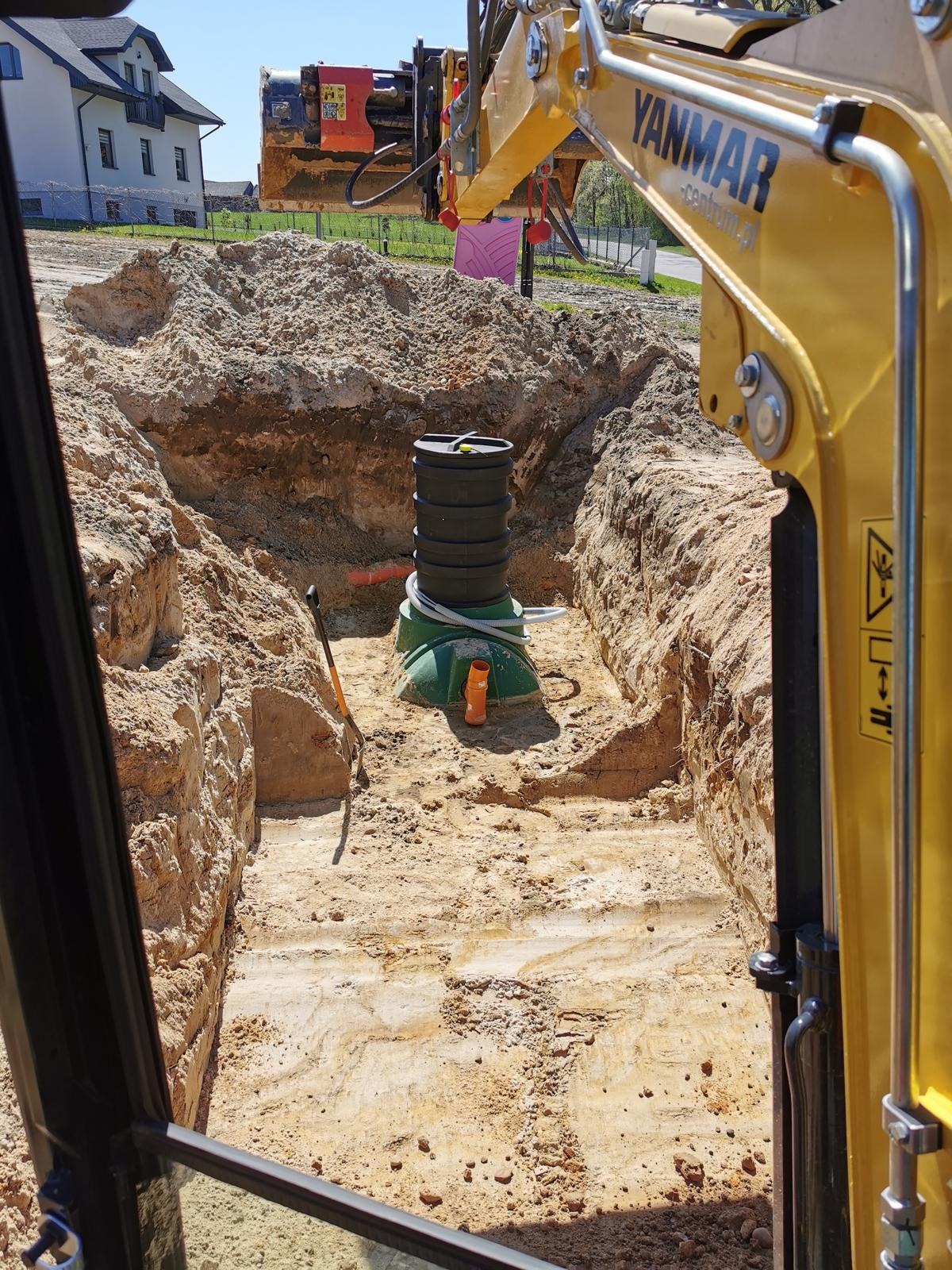Montaż przydomowej oczyszczalni ścieków w wykopie, widok z kabiny koparki Yanmar, z elementami instalacji i narzędziami. W tle budynek mieszkalny.