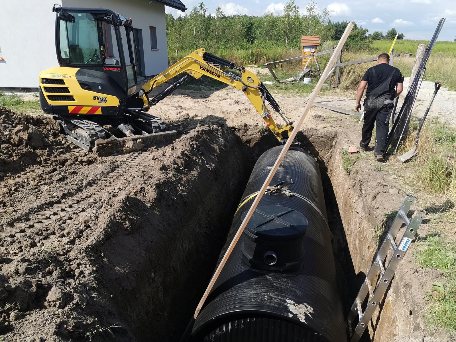 Żółta mini koparka Yanmar SV26 obok wykopu z czarnym zbiornikiem. W tle budynek i zieleń. Prace ziemne w toku, widoczna drabina i pracownik w oddali.