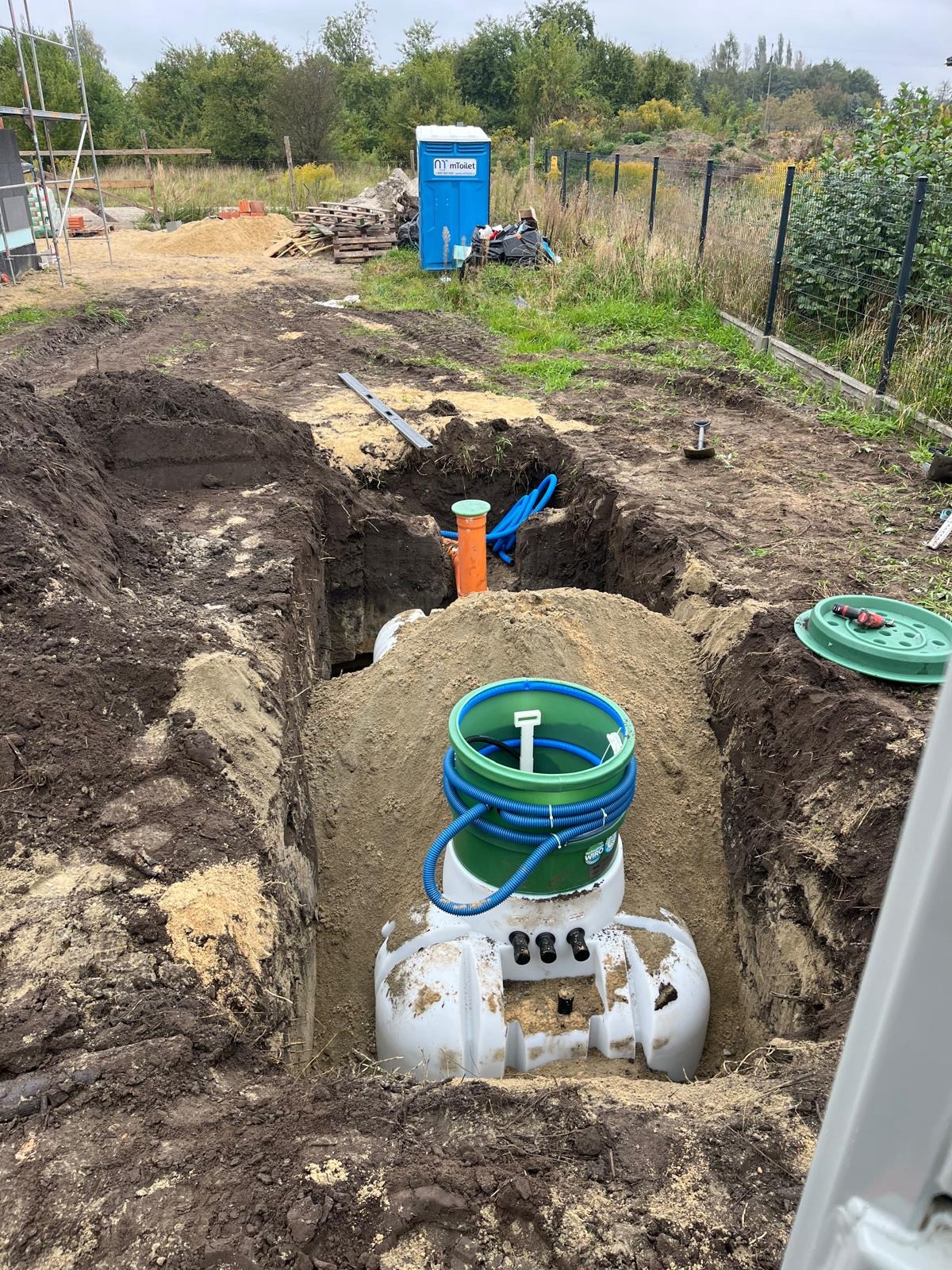 Montaż przydomowej oczyszczalni: biały zbiornik z zielonymi elementami w wykopie, niebieskie rury, piasek, ziemia. W tle TOI TOI i zieleń.