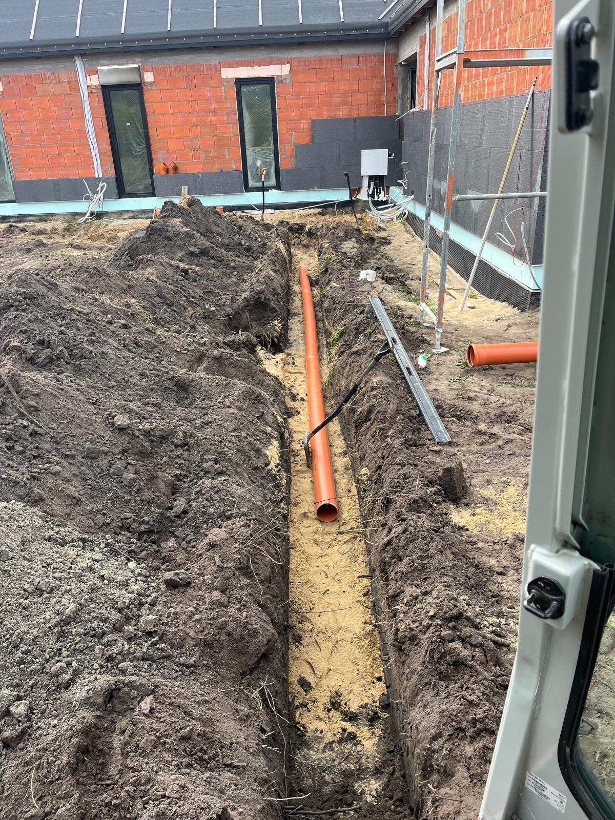 Pomarańczowa rura ułożona w wykopie z piaskowym podsypem, widok z kabiny maszyny. W tle budynek w trakcie budowy z oknami i ociepleniem.