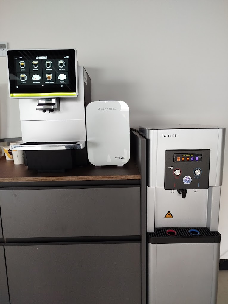 Nowoczesny ekspres do kawy z panelem dotykowym obok dystrybutora wody Ruhens i mini lodówki Venezia, ustawione na blacie szafki w biurze.