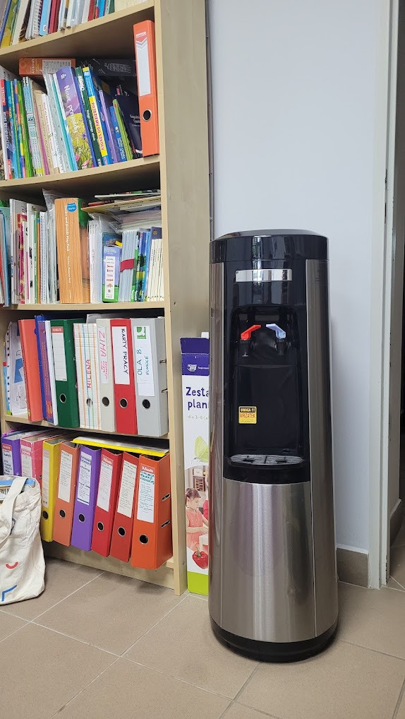 Srebrny dystrybutor wody z czerwonym i niebieskim kranikiem, stojący obok regału z książkami i segregatorami w biurze. Widoczny karton z napisem 'Zesta plan'.