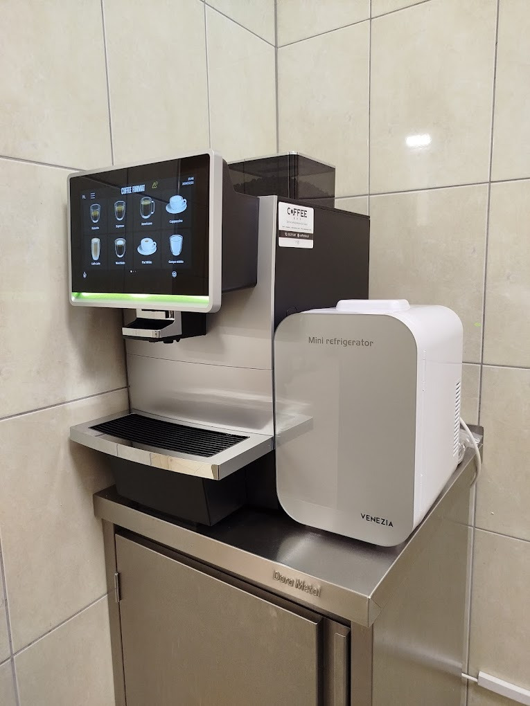 Profesjonalny ekspres do kawy z mini lodówką Venezia, umieszczony na metalowej szafce. Ekran dotykowy z wyborem napojów. Nowoczesny design w jasnej przestrzeni.