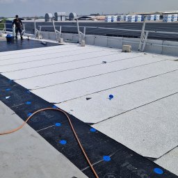 M.O.K.X-Construct Kuba Mazur - Białe pasy papy termozgrzewalnej układane na czarnej membranie z niebieskimi mocowaniami na dachu przemysłowym. W tle widoczny pracownik i budynki.