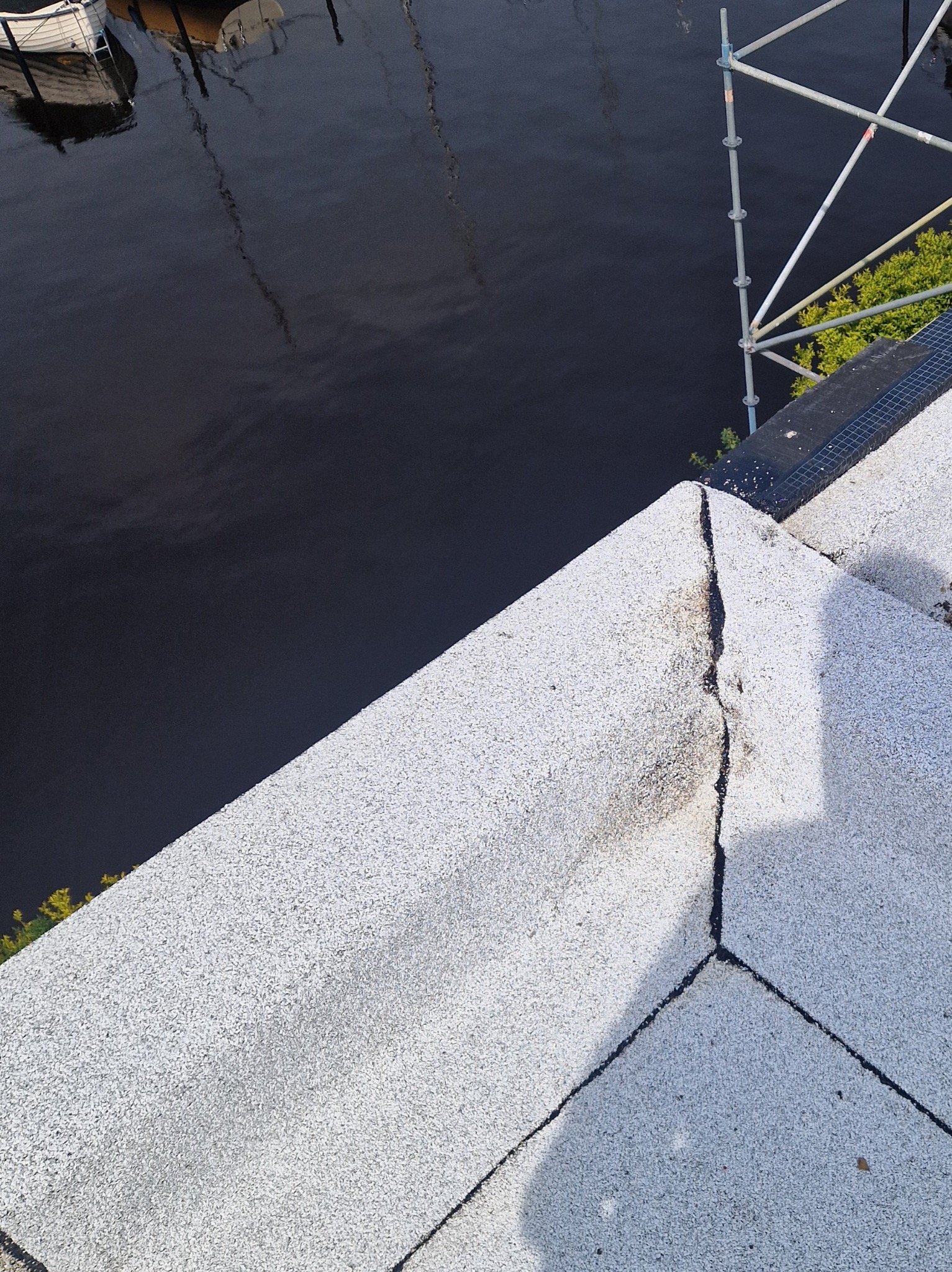 Uszkodzony, płaski dach pokryty jasnym żwirem, z widocznym pęknięciem na łączeniu. W tle ciemna woda i fragment rusztowania.