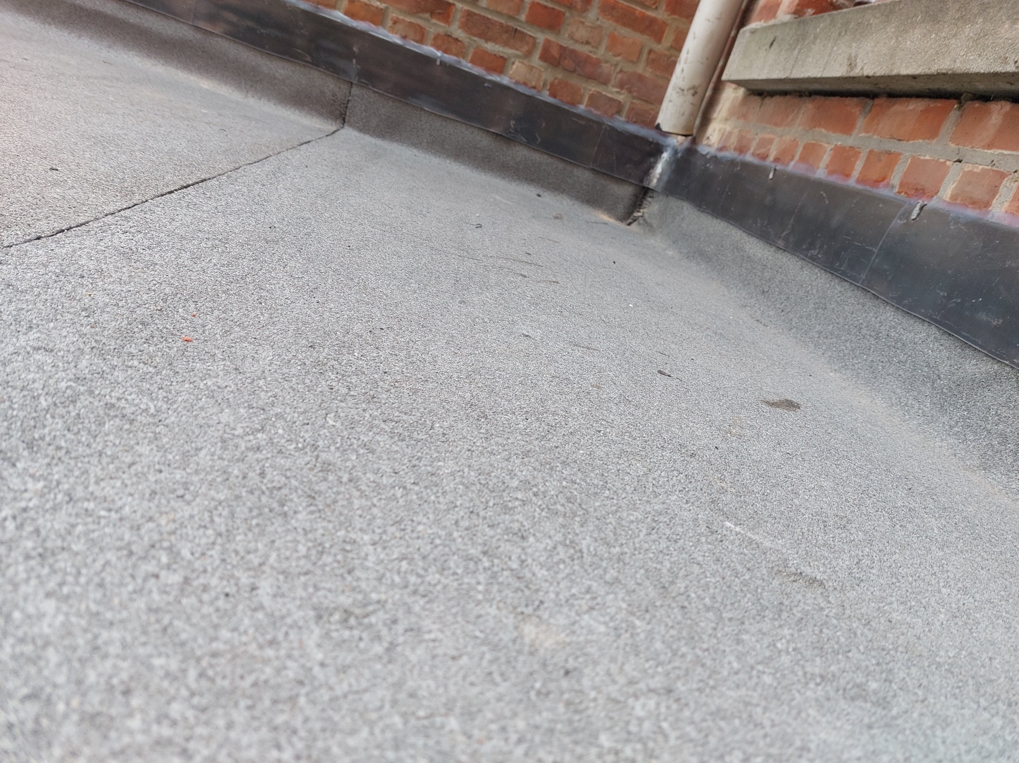 Szary, płaski dach pokryty papą, z widocznym fragmentem muru z czerwonej cegły i rynną spustową. Szczegółowe ujęcie na hydroizolację dachu.
