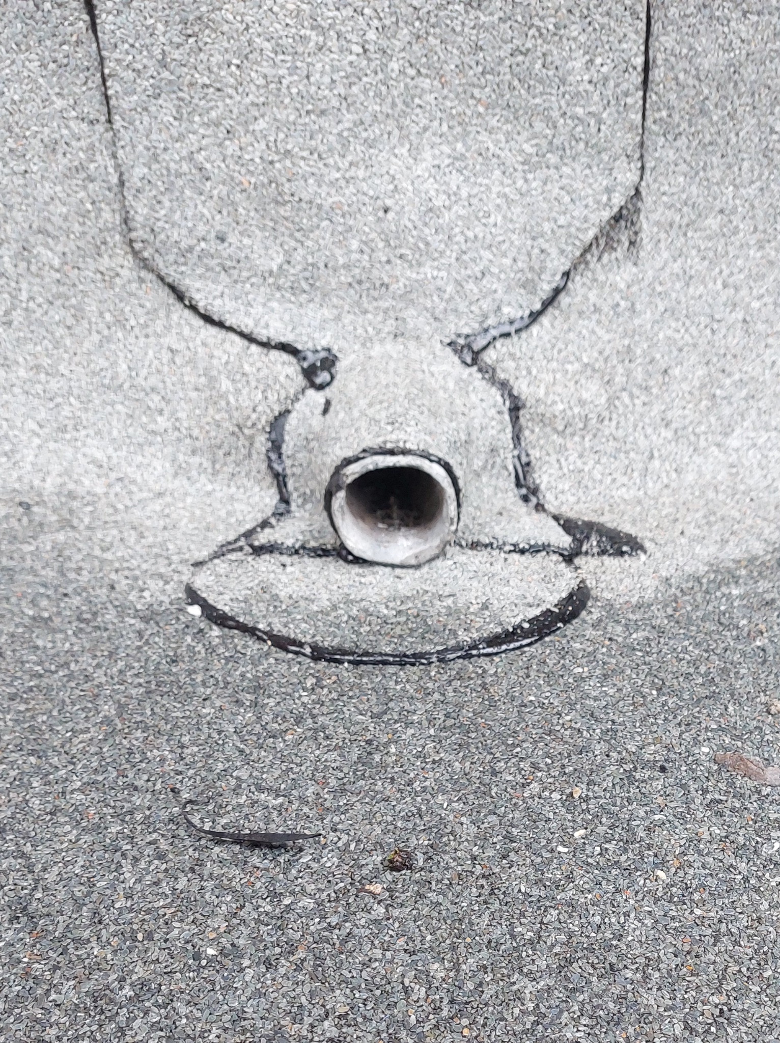 Szczegółowe ujęcie odpływu na dachu pokrytym szarym gontem bitumicznym, uszczelnionego czarnym materiałem. Widoczny fragment dachu z drobnymi kamyczkami.