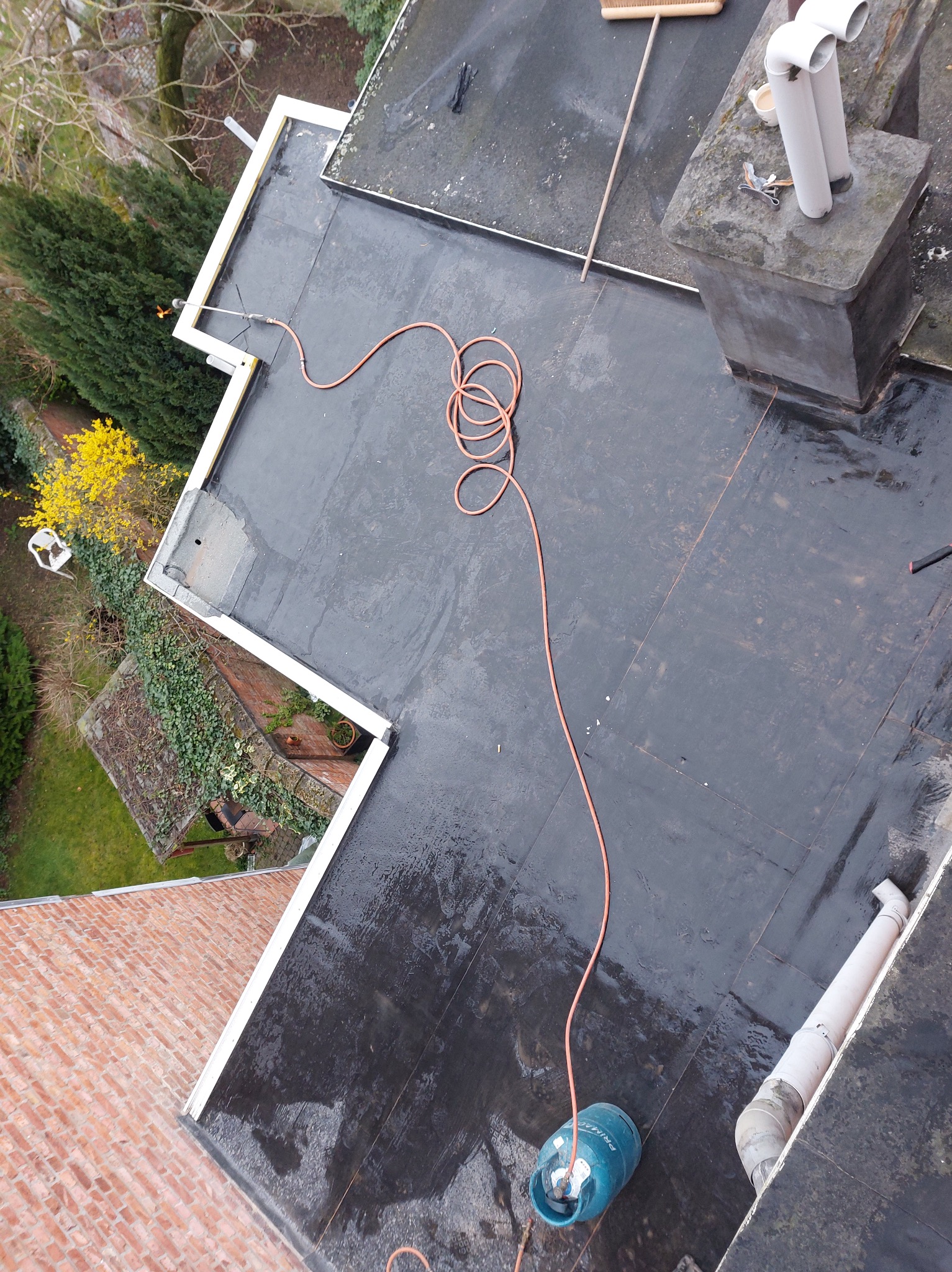 Widok z góry na płaski, czarny dach pokryty papą, z butlą gazową, wężem i narzędziami. Widoczne kominy, rynny i fragmenty otoczenia z zielenią.