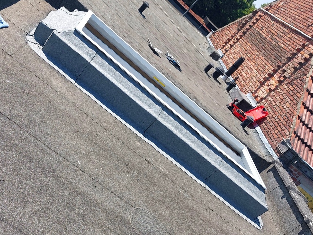 Płaski dach pokryty papą z obróbką świetlika dachowego. Widoczne narzędzia dekarskie i fragment dachu z dachówki ceramicznej w tle. Ujęcie z góry.