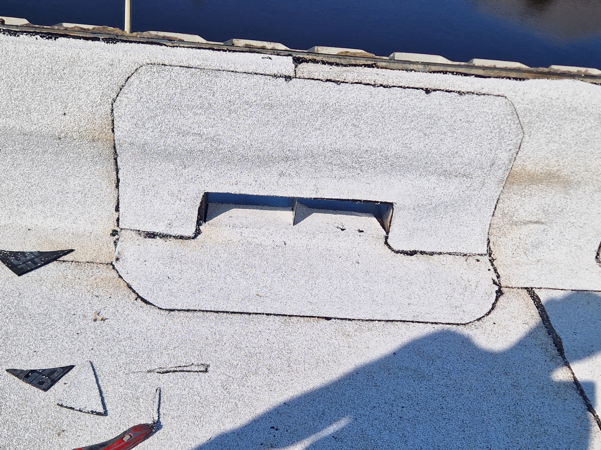 Biały dach pokryty papą z wyciętym otworem wentylacyjnym. Widoczne narzędzie dekarskie i fragment cienia. Ujęcie z góry, ostre światło słoneczne.