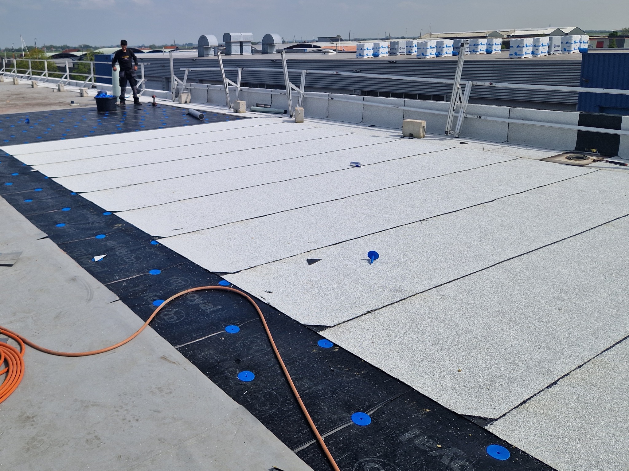 Białe pasy papy termozgrzewalnej układane na czarnej membranie z niebieskimi mocowaniami na dachu przemysłowym. W tle widoczny pracownik i budynki.