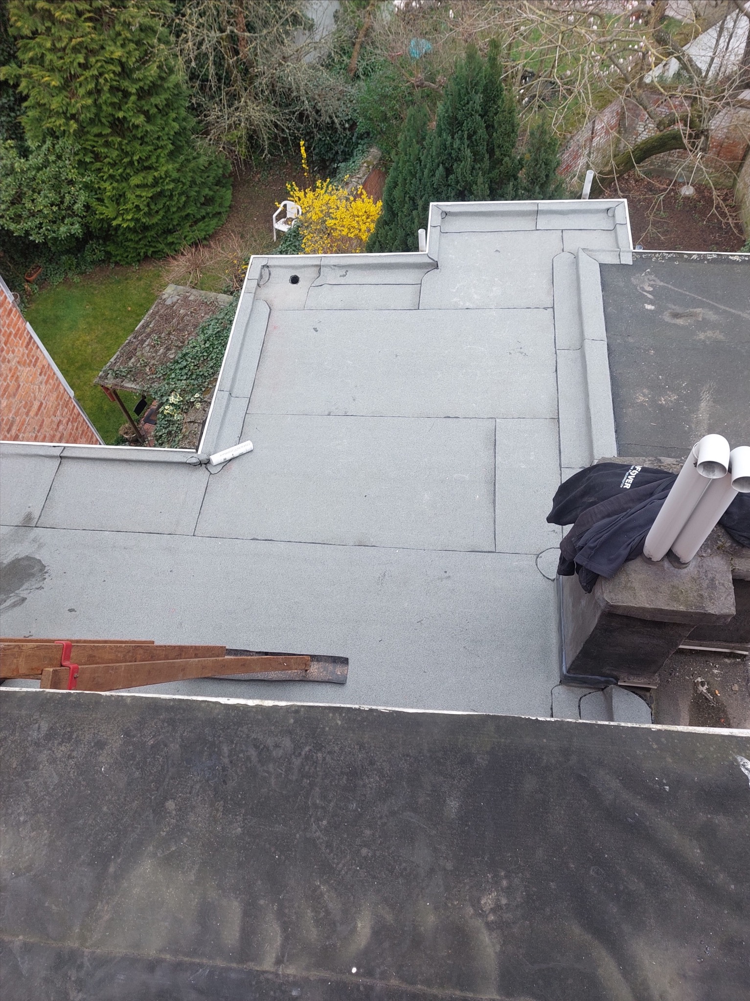 Szary, płaski dach pokryty papą termozgrzewalną z kominem i elementami wentylacyjnymi. Widoczne narzędzia dekarskie i fragment ogrodu w tle.