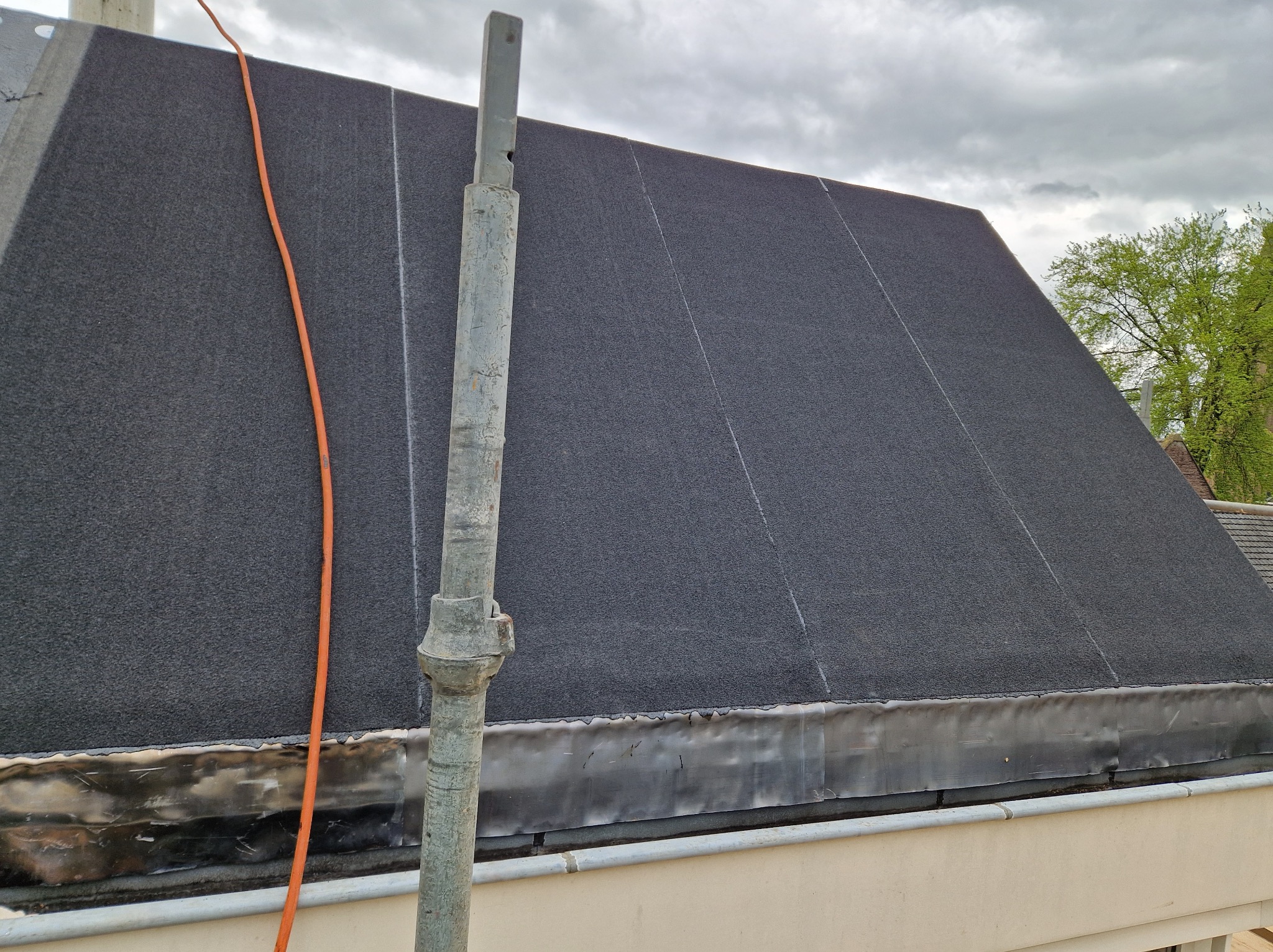 Czarny, płaski dach pokryty papą termozgrzewalną z widoczną obróbką blacharską, pionowy przewód kominowy i pomarańczowy kabel na tle pochmurnego nieba.