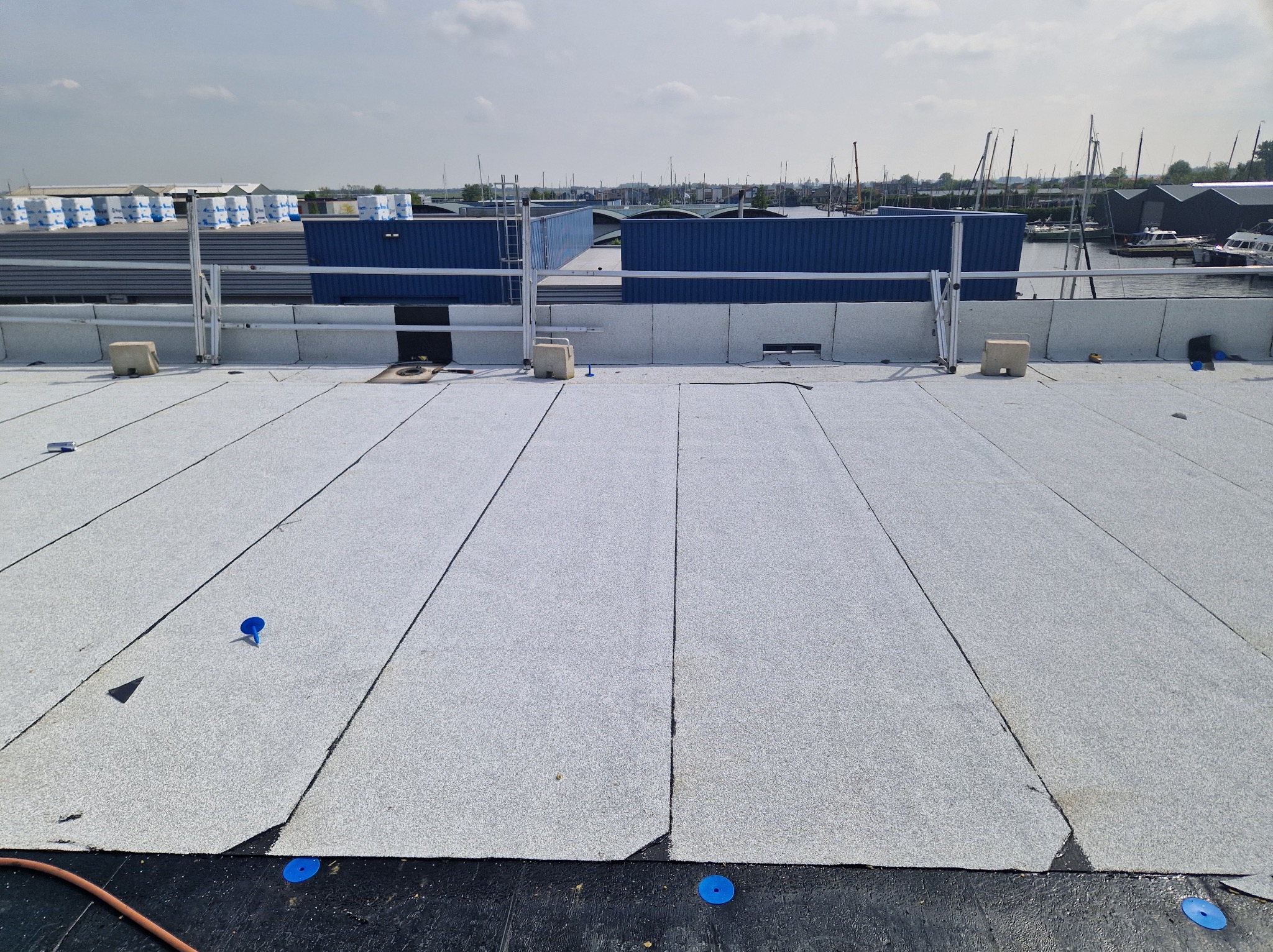 Szary dach płaski z widocznymi łączeniami papy i niebieskimi elementami mocującymi. W tle budynki przemysłowe i marina z jachtami w słoneczny dzień.