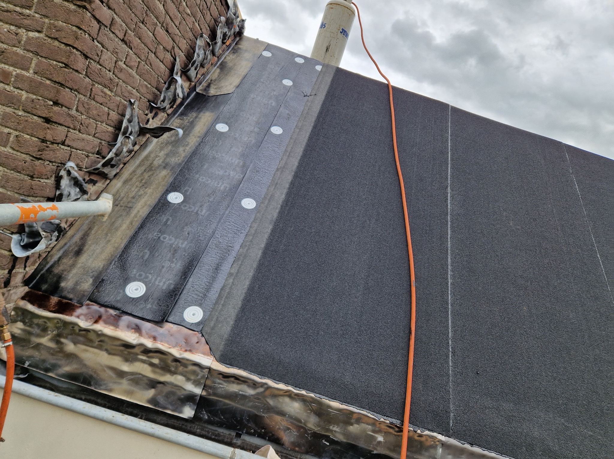 Fragment dachu z nową, czarną papą bitumiczną, mocowaną mechanicznie. Widoczne detale łączeń i obróbek blacharskich przy kominie i ścianie. Pomarańczowy kabel na dachu.