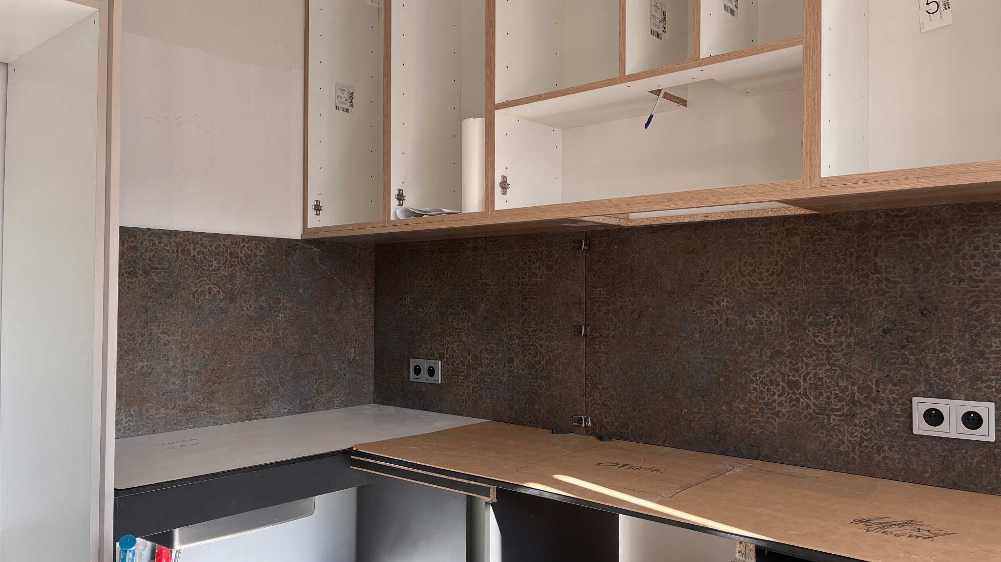 Montaż szafek kuchennych z brązową mozaiką na ścianie. Widoczne gniazdka elektryczne i elementy mocujące. Etap prac wykończeniowych w kuchni.