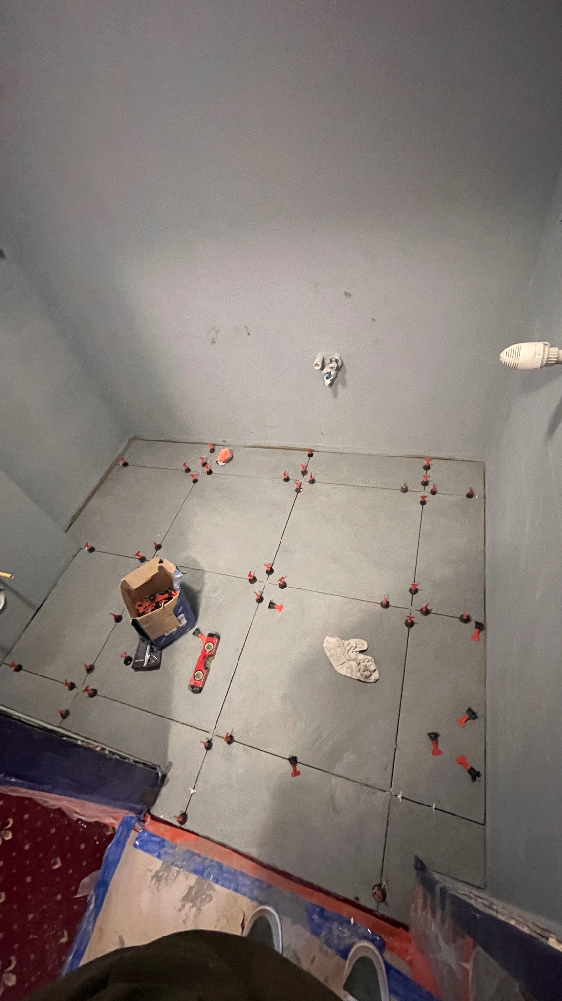 Szare płytki podłogowe w trakcie układania, z systemem poziomowania, widoczne narzędzia i karton z elementami montażowymi. Prace glazurnicze w łazience.