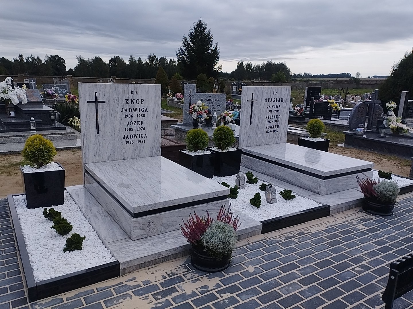 Dwa marmurowe nagrobki z wygrawerowanymi napisami i krzyżami, otoczone białymi kamykami i roślinami w donicach, na tle cmentarza i pochmurnego nieba.