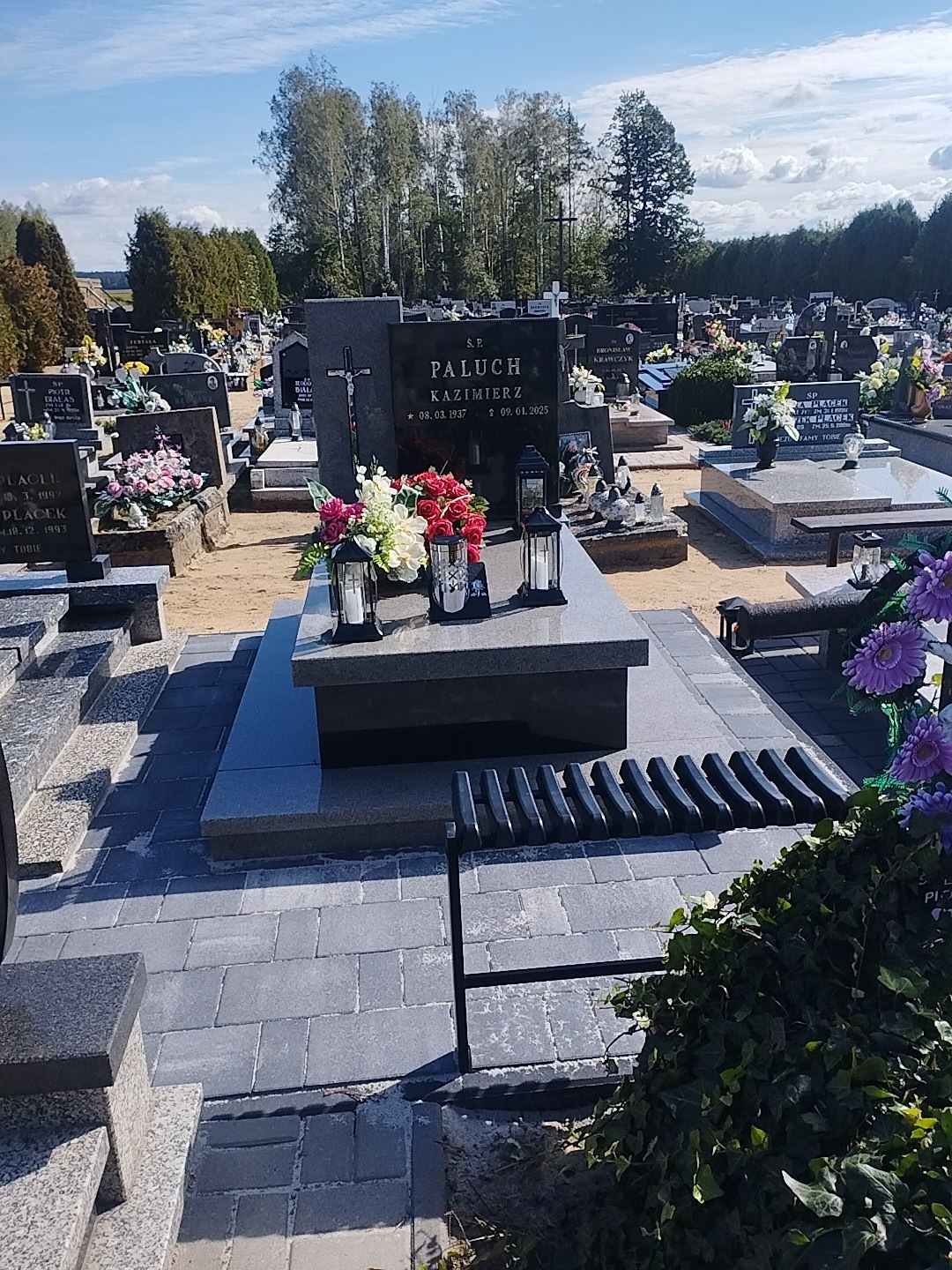 Granitowy nagrobek z kwiatami i lampionami na cmentarzu, otoczony kostką brukową. W tle drzewa i inne groby. Widok w słoneczny dzień.