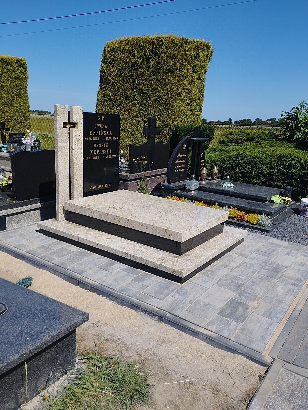 Jasny nagrobek z krzyżem na cmentarzu, otoczony kostką brukową i zielenią. Widoczne inne groby w tle, błękitne niebo. Ujęcie z poziomu gruntu.