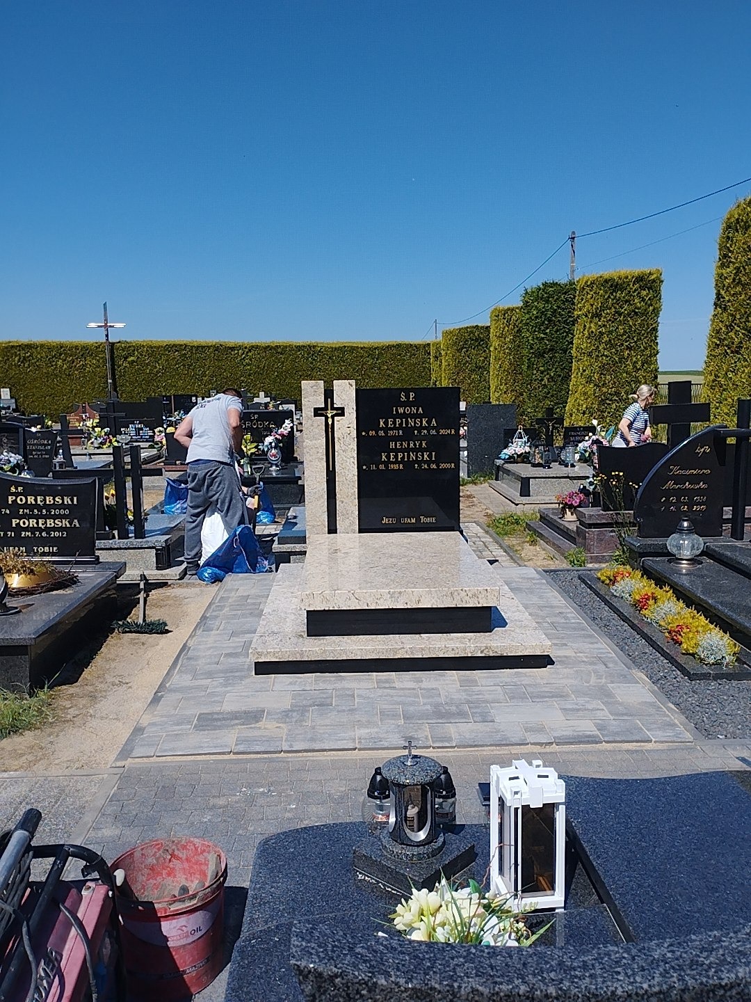 Granitowy nagrobek z krzyżem na cmentarzu, otoczony kostką brukową. Widoczna osoba sprzątająca. W tle żywopłot i inne groby. Słoneczny dzień.