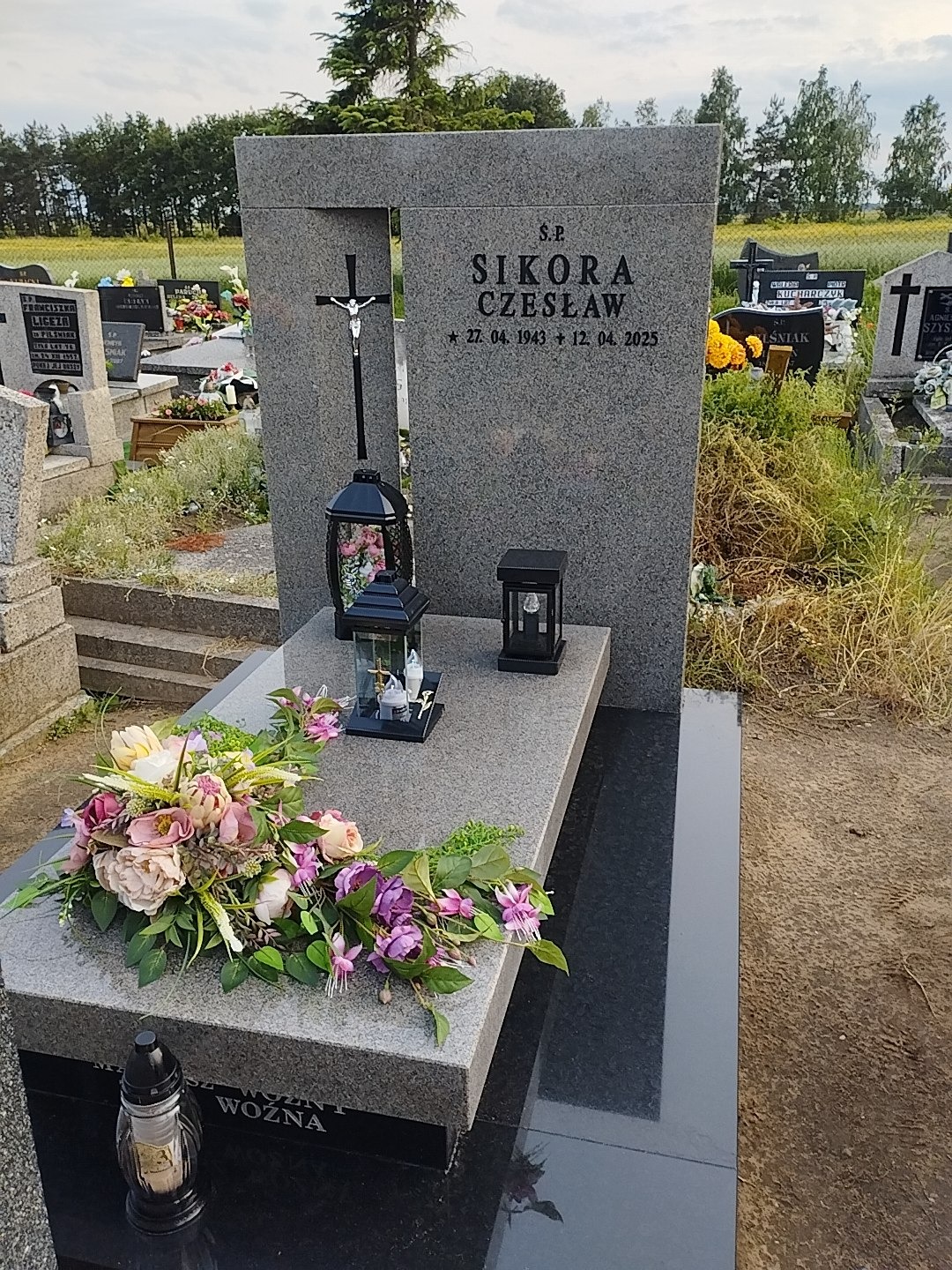 Granitowy nagrobek z krzyżem i inskrypcją 'S.P. Sikora Czesław' otoczony kwiatami i lampionami na cmentarzu, w tle zieleń drzew i inne groby.