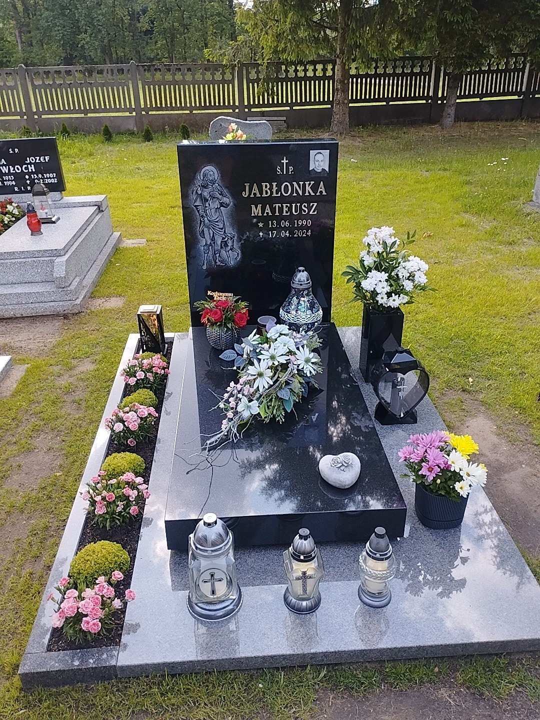 Czarny nagrobek z jasnoszarego granitu, zdobiony kwiatami i lampionami. Na płycie wyryty wizerunek świętego i dane zmarłego. Otoczenie: trawa, drzewa, drewniany płot.