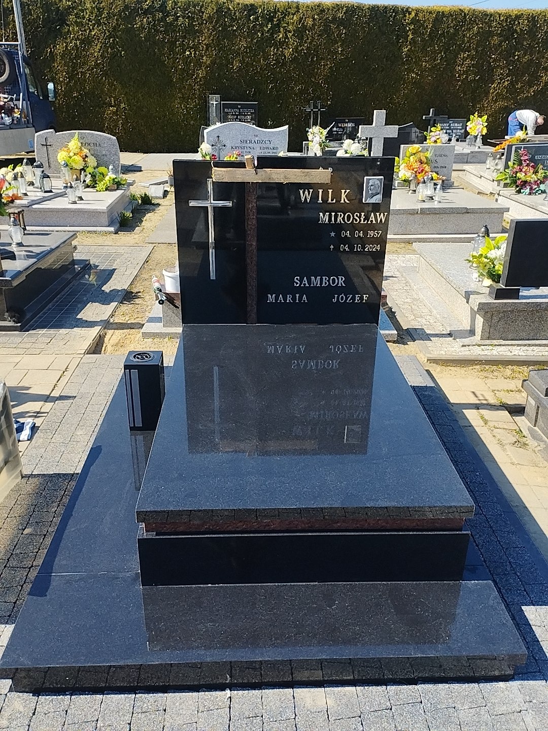 Czarny, błyszczący nagrobek z wygrawerowanym krzyżem i danymi zmarłego na tle cmentarza. Widoczne inne nagrobki i zieleń.