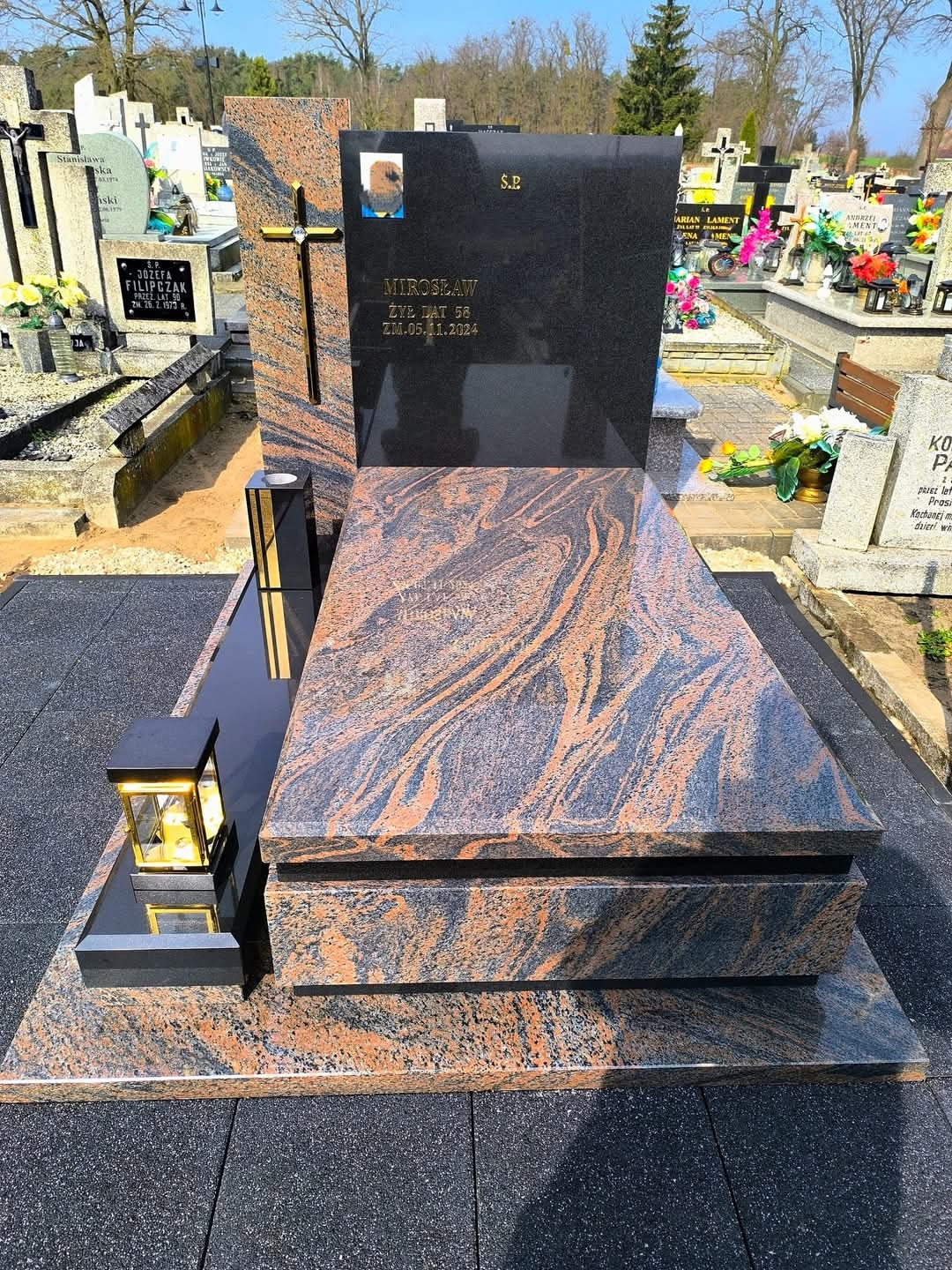 Granitowy nagrobek z krzyżem i lampionami, w odcieniach czerni i brązu, na cmentarzu. Widoczne inskrypcje i zdjęcie osoby zmarłej. Detal kamieniarski.