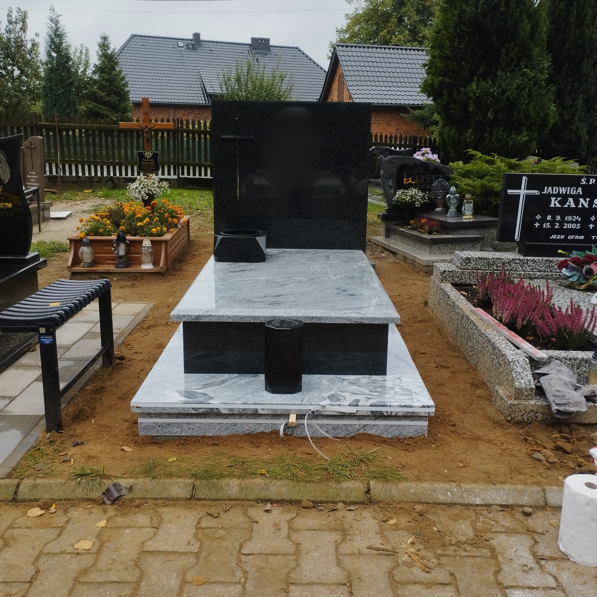 Granitowy nagrobek z czarną tablicą i szarym obramowaniem na cmentarzu. Widoczne inne groby i zieleń w tle. Minimalistyczny design z elementami dekoracyjnymi.