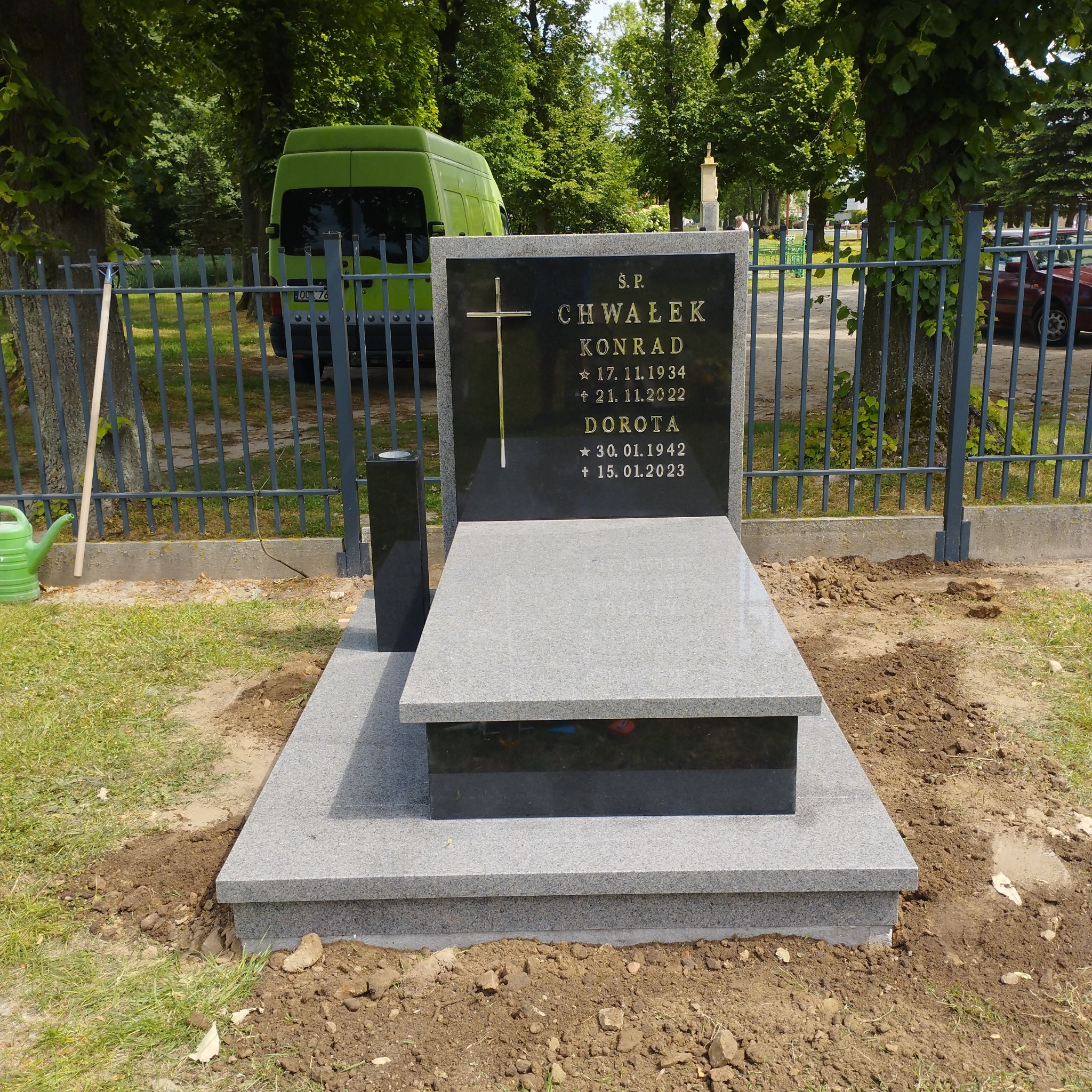 Granitowy nagrobek z czarną tablicą inskrypcyjną i złotym krzyżem, otoczony zielenią cmentarza. Widoczny fragment zielonego vana w tle, sugerujący transport.