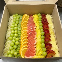 KRAKPRESS SPÓŁKA Z OGRANICZONĄ ODPOWIEDZIALNOŚCIĄ - Elegancka kartonowa taca z owocami: jabłka, winogrona, kiwi, pomarańcze, grejpfruty, mango, śliwki i ananas, ułożone w rzędach, idealne na catering.