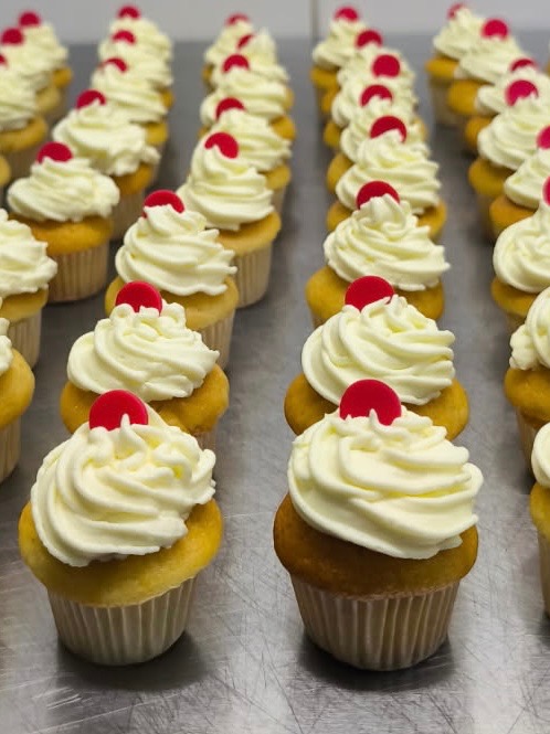 Rząd równych babeczek z kremem i czerwoną wisienką na wierzchu, ustawionych na metalowej powierzchni. Idealne na słodki poczęstunek podczas imprezy firmowej.