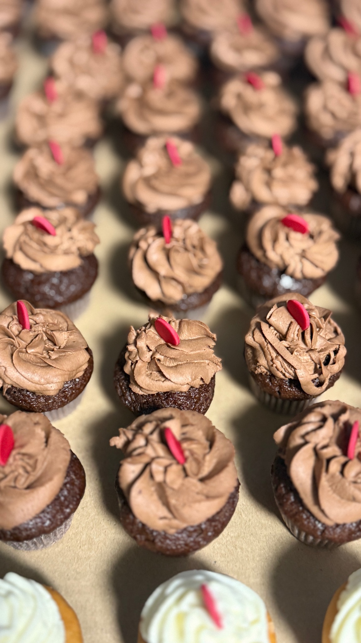 Rząd czekoladowych babeczek z kremem i czerwoną dekoracją, ułożonych na beżowym tle. Część babeczek z białym kremem widoczna w dolnej części kadru.