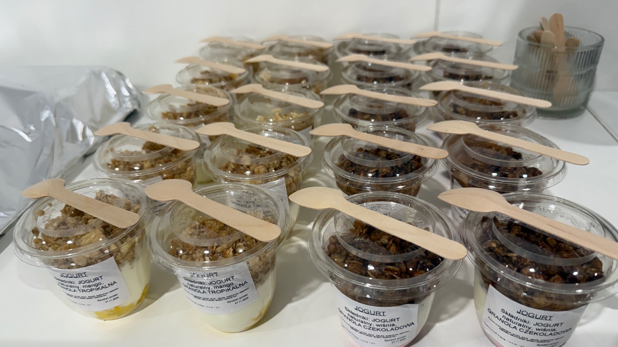 Rząd plastikowych pojemników z jogurtem i granolą, każdy z drewnianą łyżeczką na wierzchu. Widoczne różne smaki: mango i czekolada z wiśnią.
