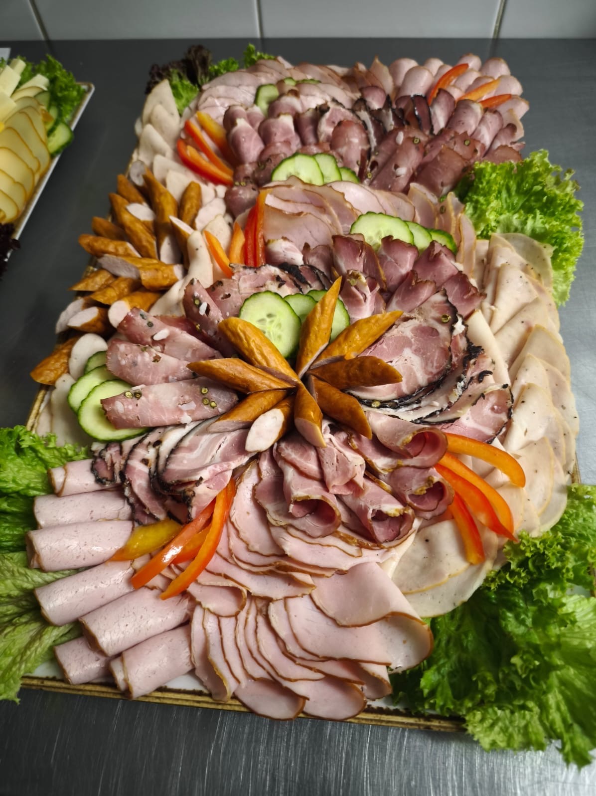 Bogato zdobiona deska wędlin i serów z dodatkiem warzyw, ułożona na metalowym stole. Widoczne różne rodzaje wędlin, kiełbas, ogórki i papryka. Idealna na catering.