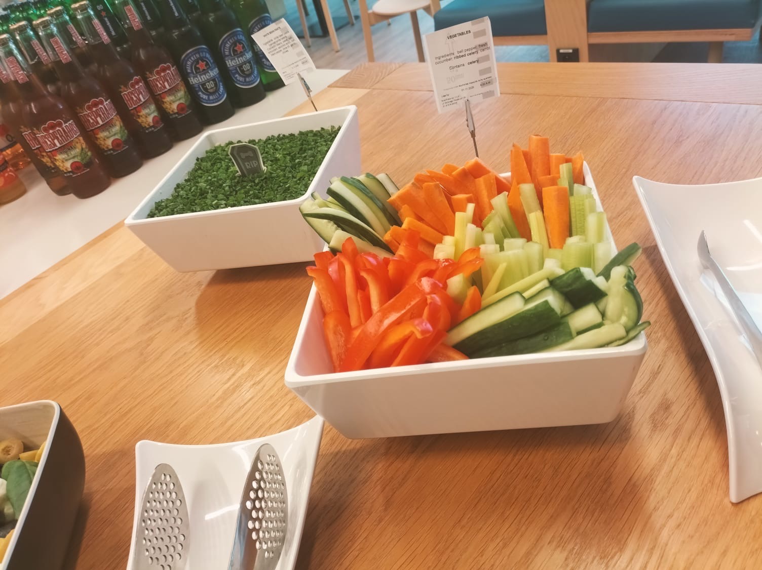 Dekoracyjnie ułożone warzywa (papryka, ogórek, marchew, seler) w białych miseczkach na drewnianym stole, obok napoje w butelkach i szczypiorek z dekoracją.