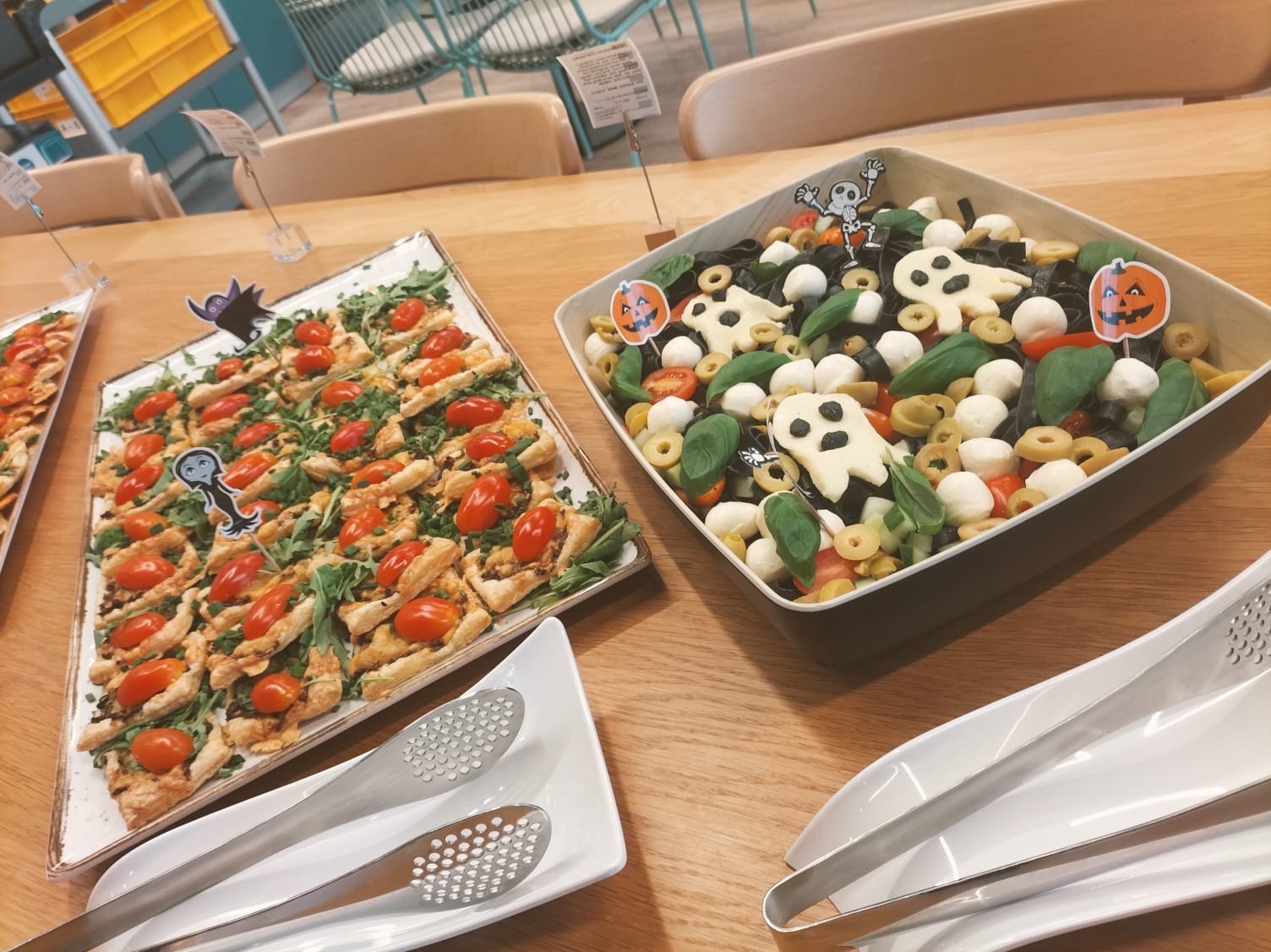 Halloweenowy catering: sałatka caprese z czarnym makaronem i mozzarella w kształcie duszków, obok tarta z pomidorami i rukolą. Bufet na drewnianym stole.